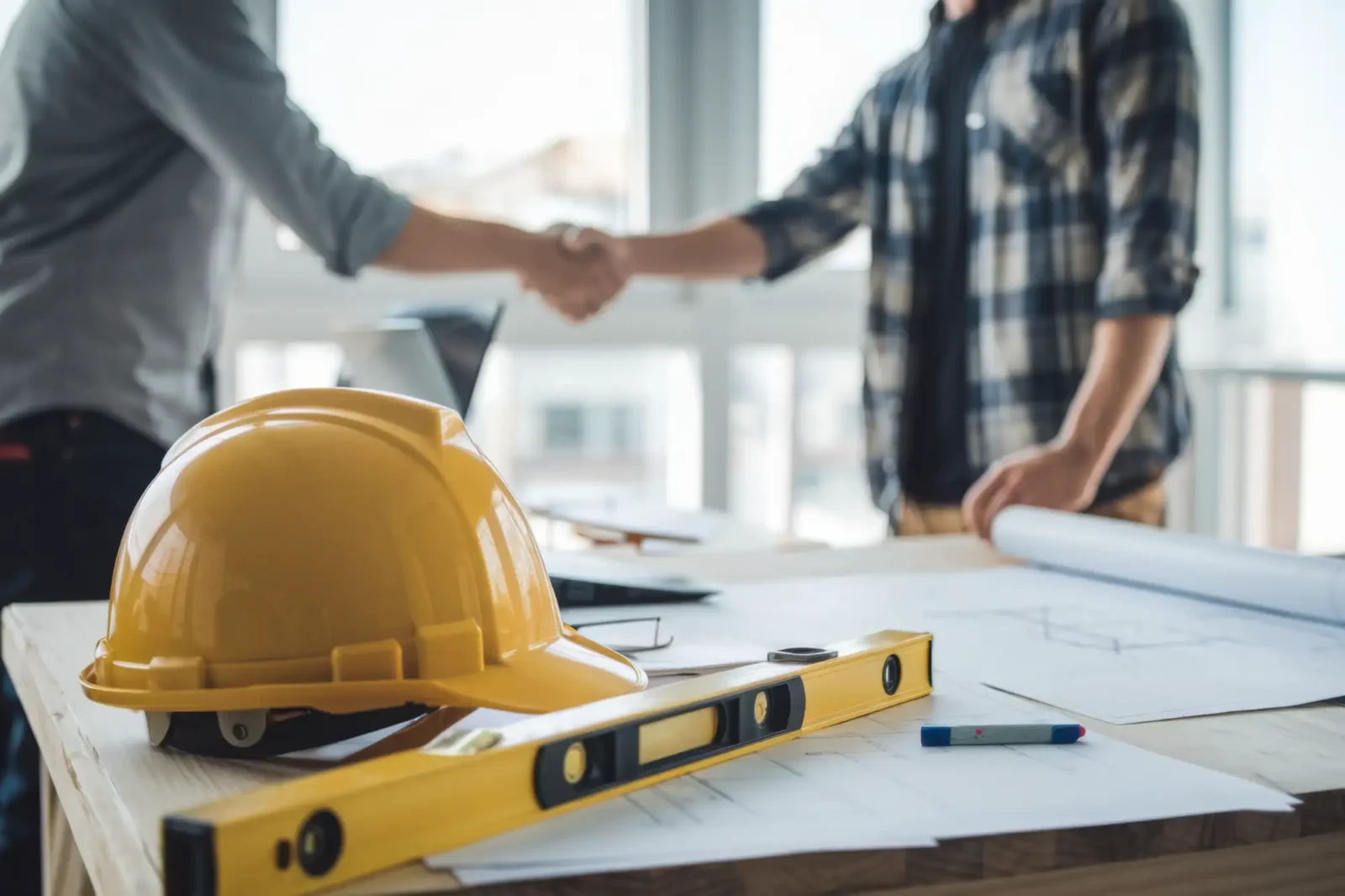 construction hat and level on a blueprint with worker and client shaking hands