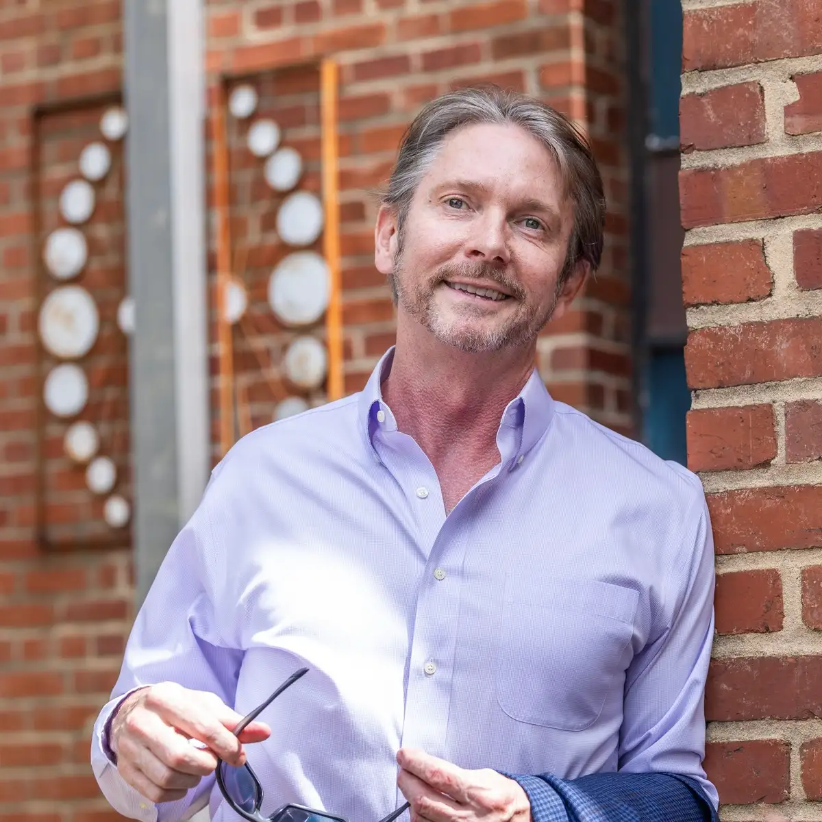 DM Management owner David McAnulty, a middle-aged smiling man wearing a lavender button up shirt and holding sunglasses