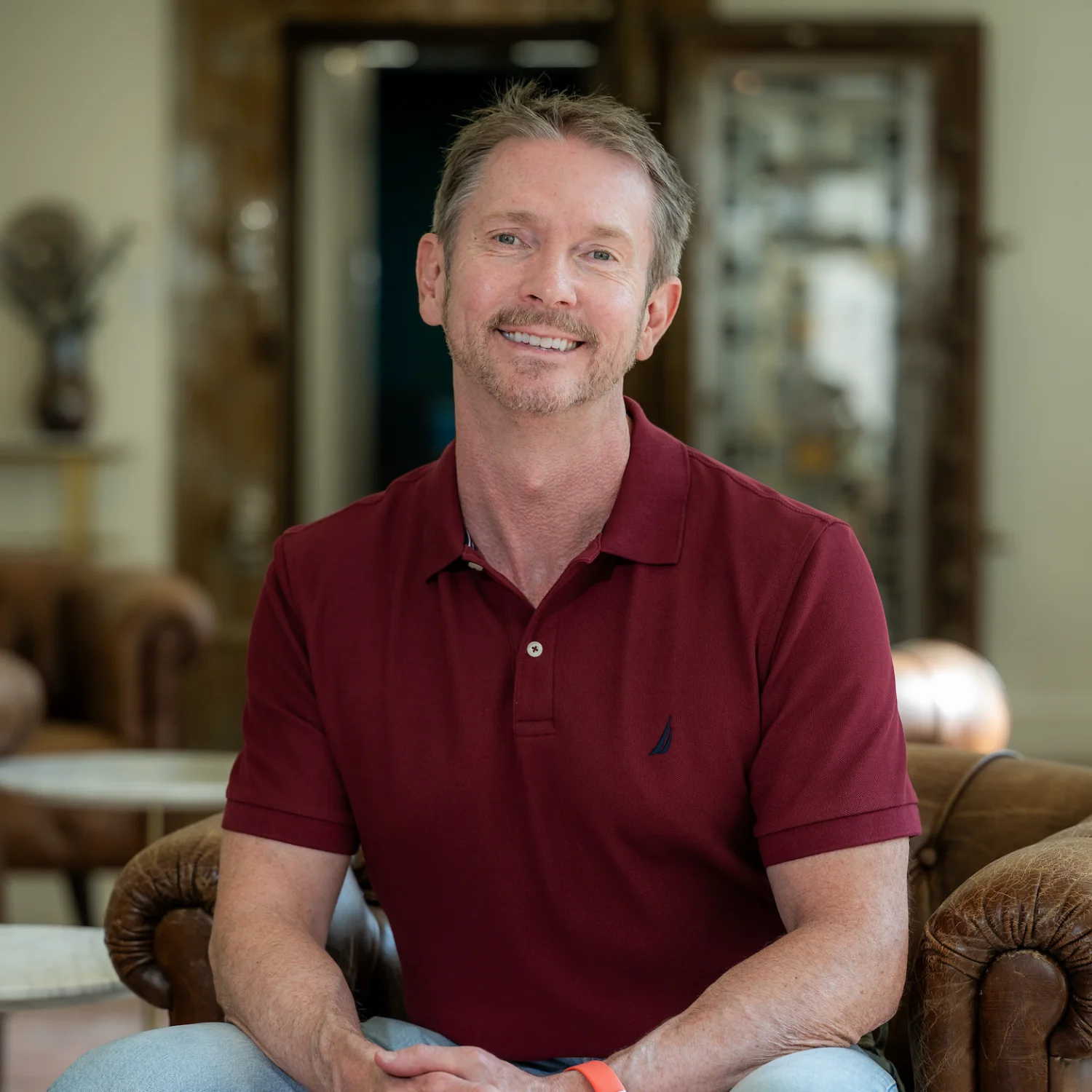 David McAnulty, wearing a dark red polo shirt, sits in a brown leather chair in an office setting, smiling directly at the viewer.