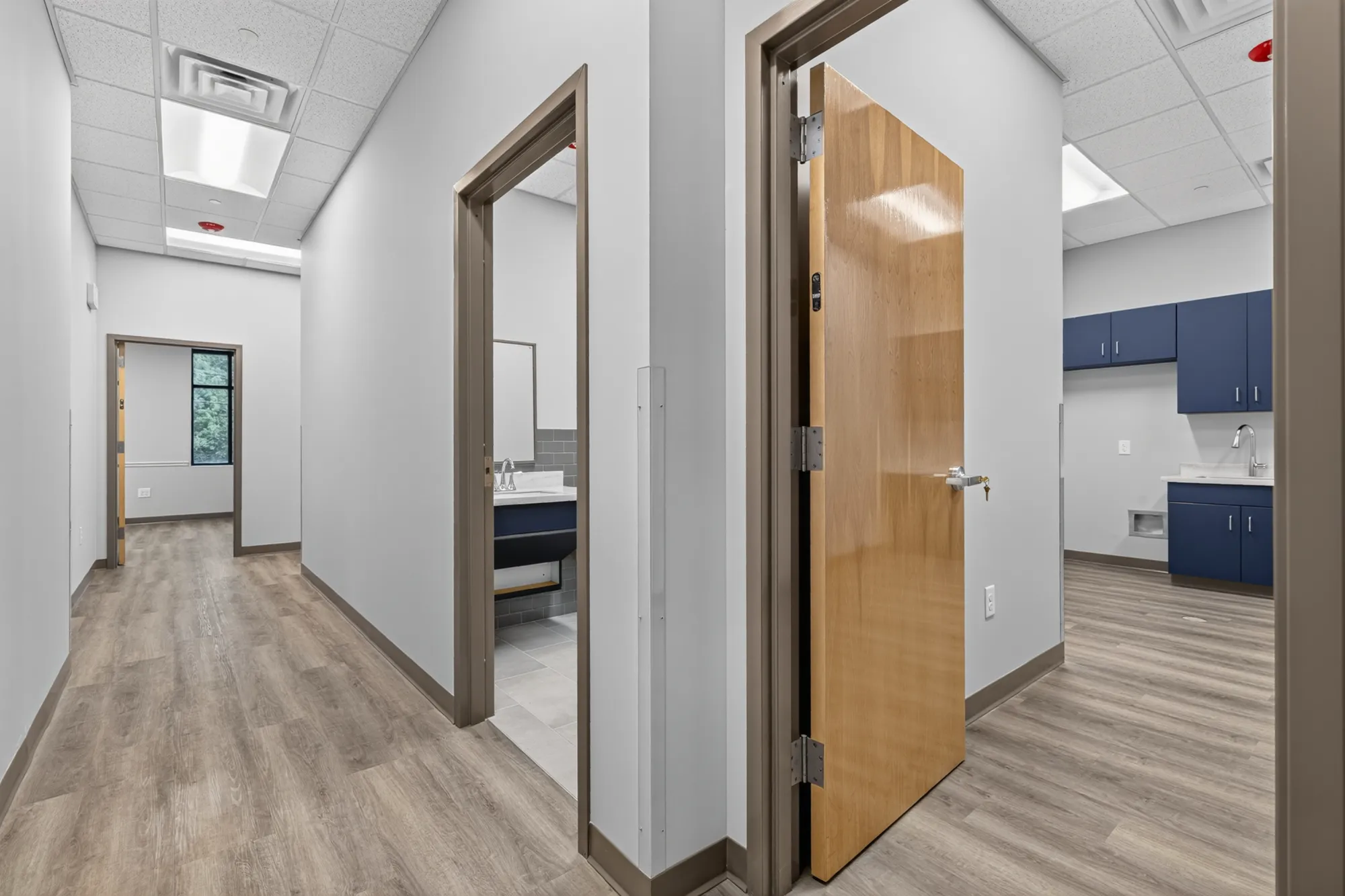 A bright, modern office hallway with wood-look flooring, white walls, and multiple doorways leading to a restroom and a lab or clinical sink area with dark blue cabinets.