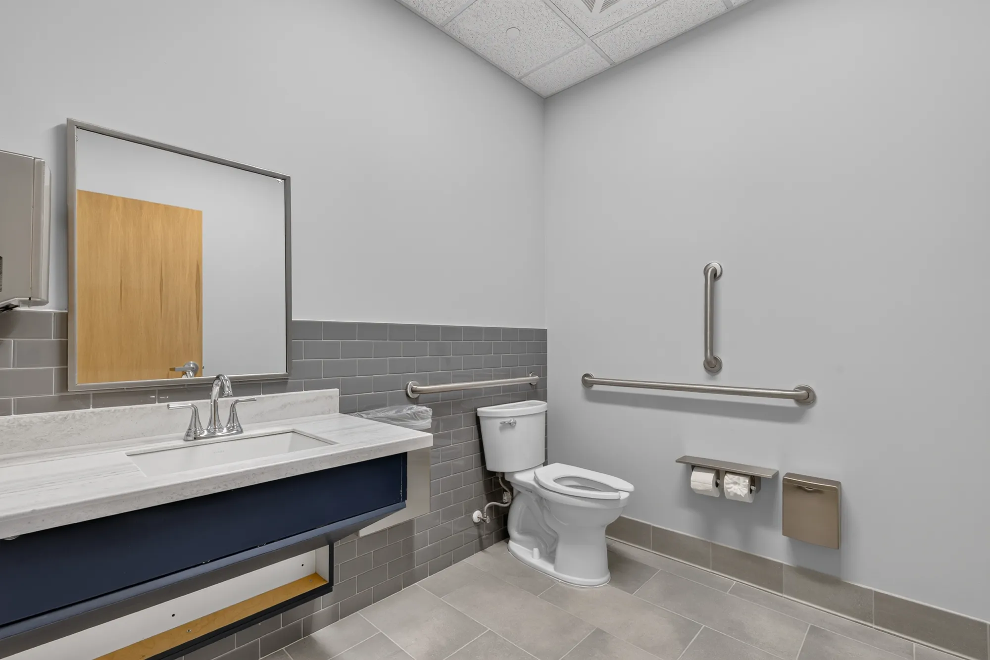 A clean, modern accessible restroom featuring a white toilet, ADA grab bars, and a floating sink with a white marble counter and dark blue vanity base, set against gray tile.