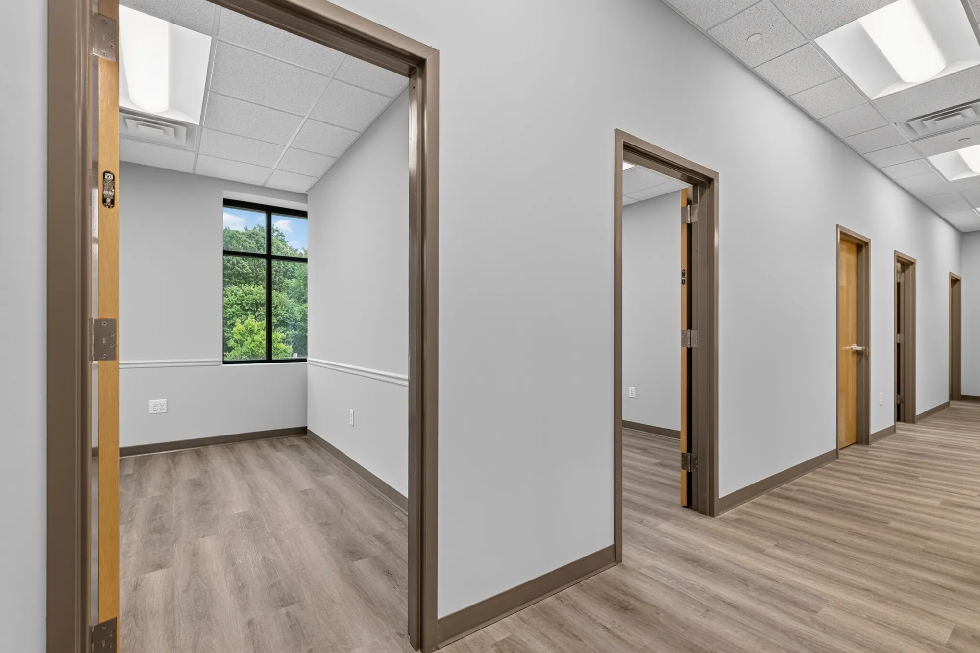 A newly constructed commercial office suite hallway with light wood-look flooring and gray walls. Multiple door frames line the hall, leading to offices with natural light.