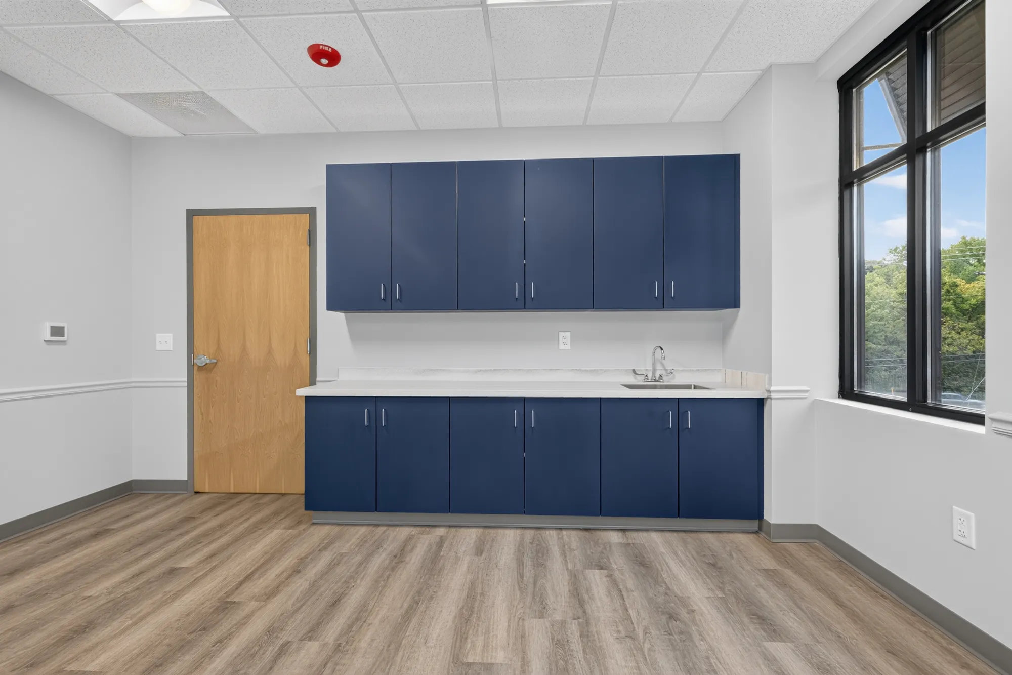 A functional room in a commercial office with navy blue cabinets, a white counter, sink, light wood door, and large window.