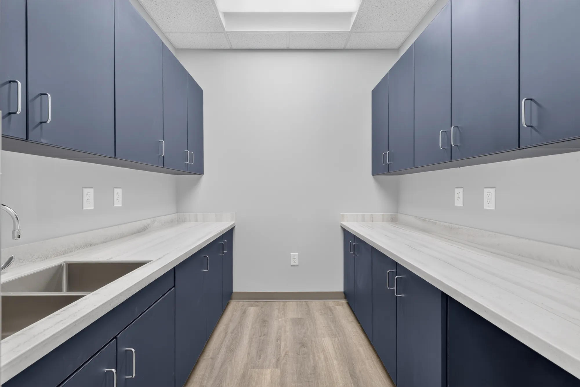 A clean, narrow medical or lab room with symmetrical dark blue cabinets and white countertops on both sides. The left counter includes a stainless steel double sink.