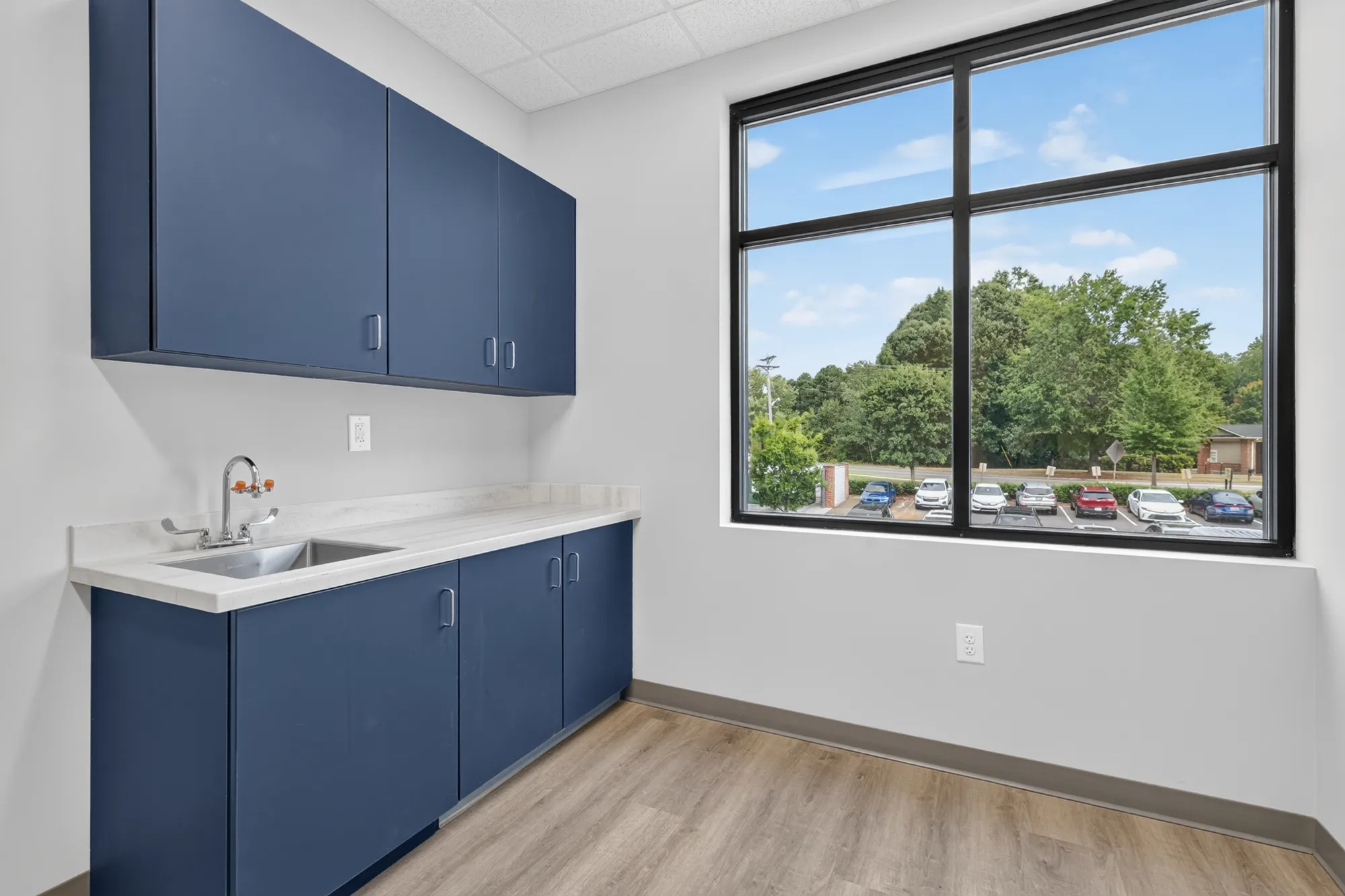 An empty room with dark blue upper and lower cabinetry, a sink, and white countertops. A large window offers a view of a parking lot and thick green trees outside.