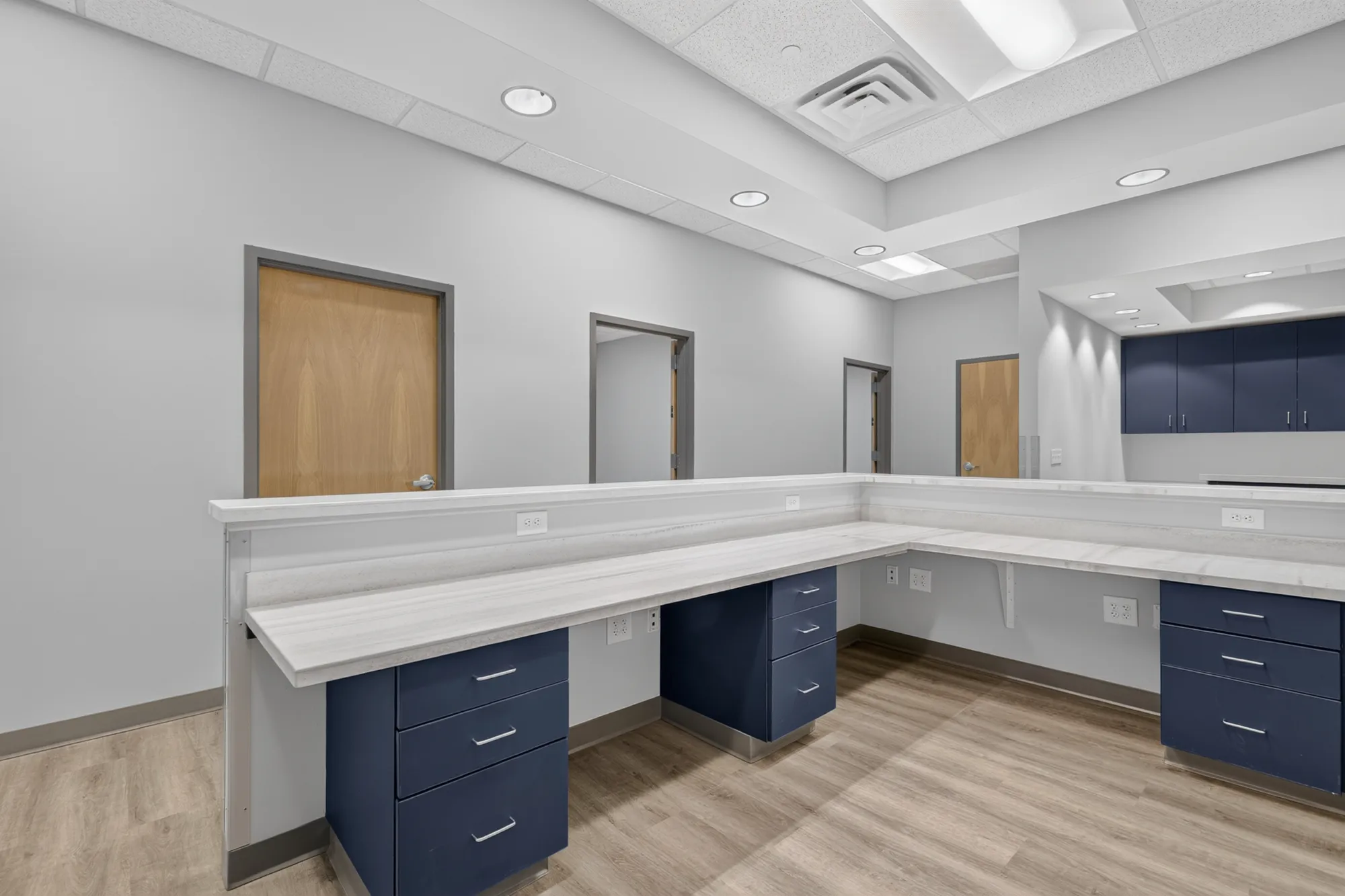 A modern administrative workspace featuring a long L-shaped reception desk with navy cabinets, light countertops, and multiple access doors along the light gray wall.