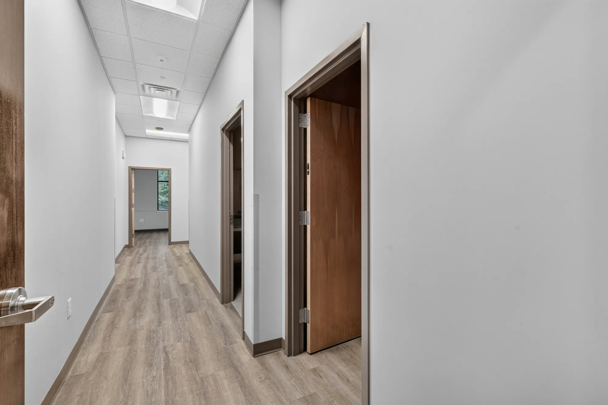 A long, brightly lit commercial hallway with light wood flooring and gray walls. Several doors line the hall; one is open, showing a room with a window to the outside.