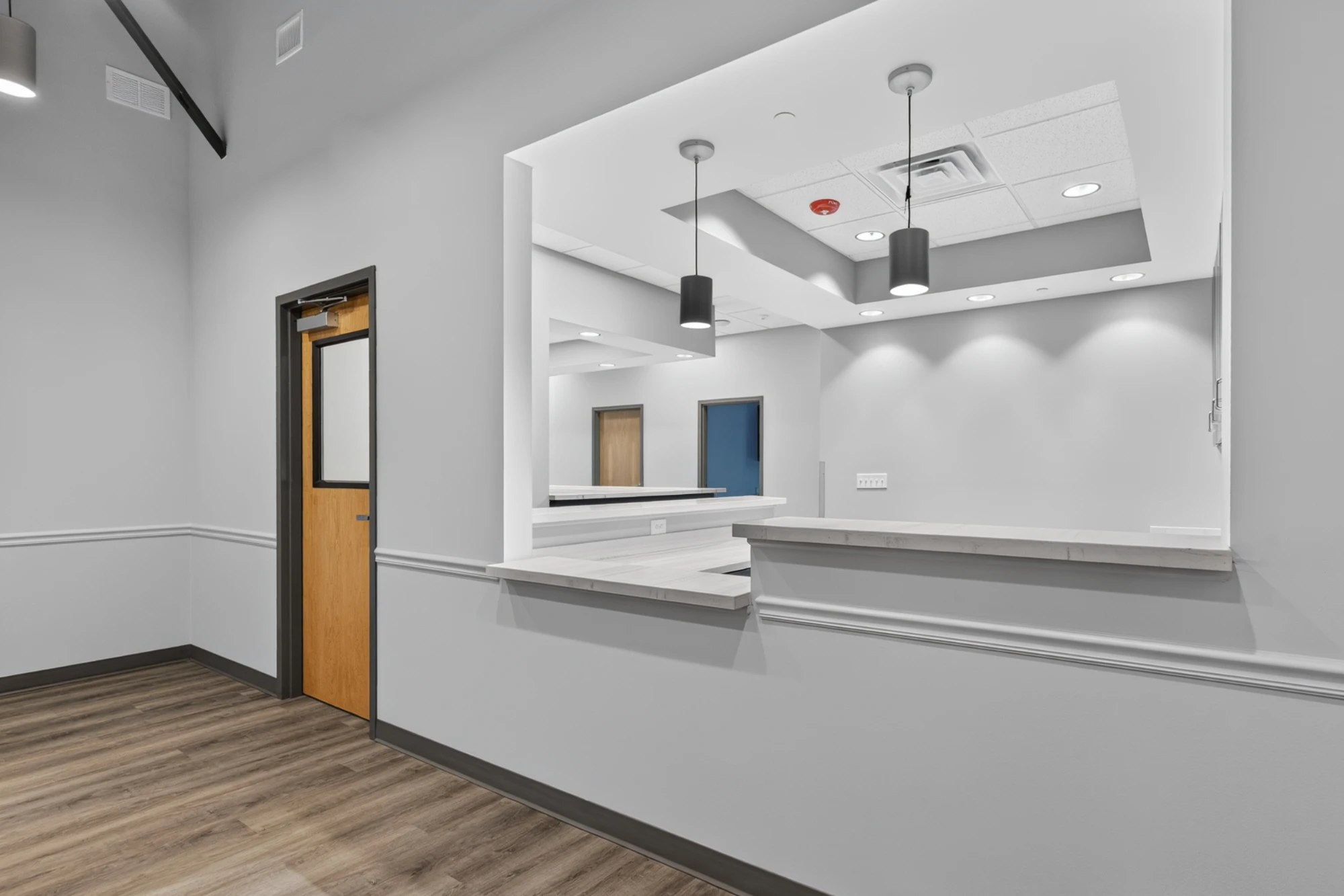 The interior view of a commercial office reception area featuring a wide service counter window and two modern pendant lights above. Light gray walls and wood flooring.