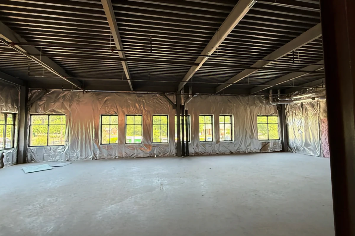 An open commercial space under construction featuring an exposed dark metal ceiling, concrete floor, and silver insulation lining the walls with multiple windows.
