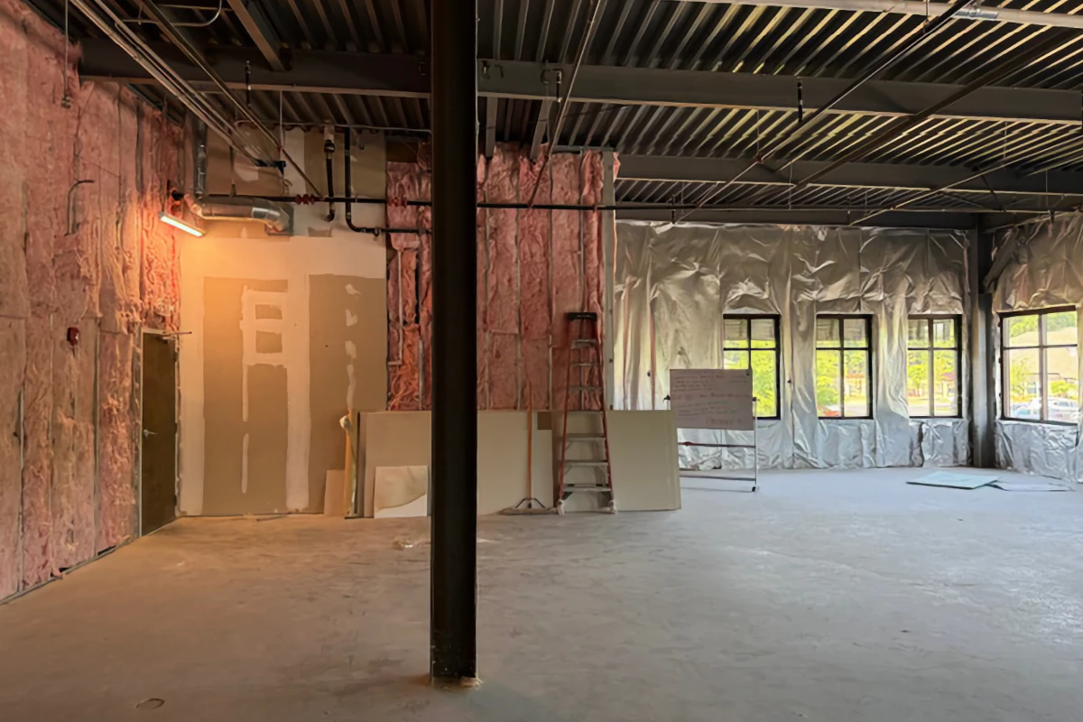 An interior view of a commercial upfit showing exposed pink wall insulation, ductwork, and new drywall. A ladder and windows are visible in the large open space.