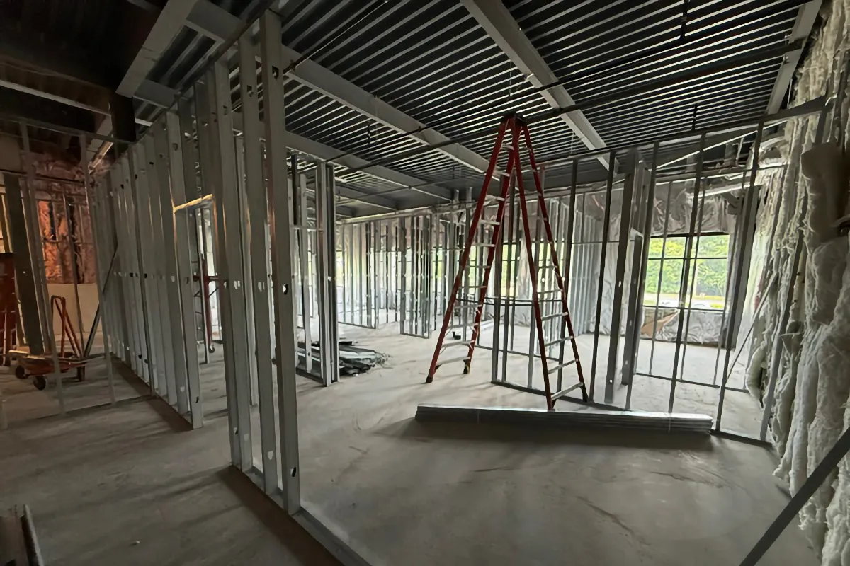 An interior view of a commercial upfit project showing the metal stud wall framing nearing completion. A red ladder stands on the concrete floor under the exposed metal deck ceiling.